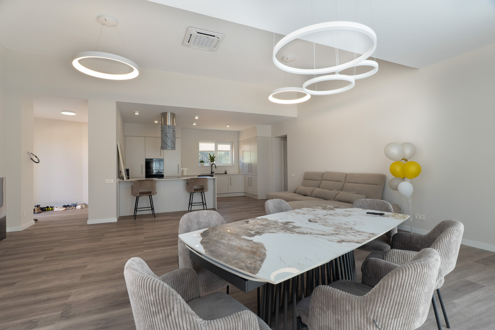 Offener Wohnbereich mit Marmortisch, gepolsterten Stühlen und minimalistischer weißer Küche mit Kochinsel sowie modernen runden LED-Pendelleuchten.