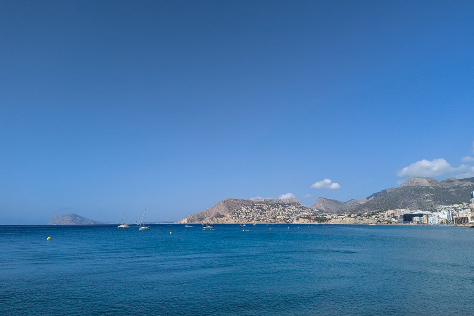 Panoramablick auf das Mittelmeer vor der Küste von Calpe mit Segelbooten und der bergigen Küstenlinie unter strahlend blauem Himmel.