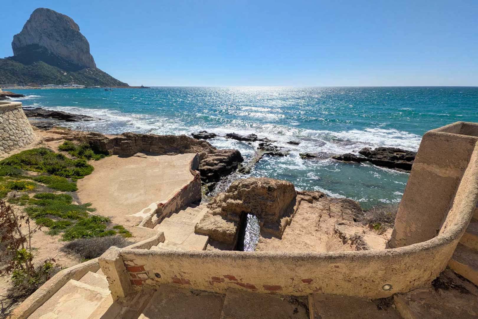 Panoramablick auf den Peñón de Ifach von einer Steinterrasse mit direktem Zugang zu einer Bucht mit türkisblauem Wasser und der Küste.