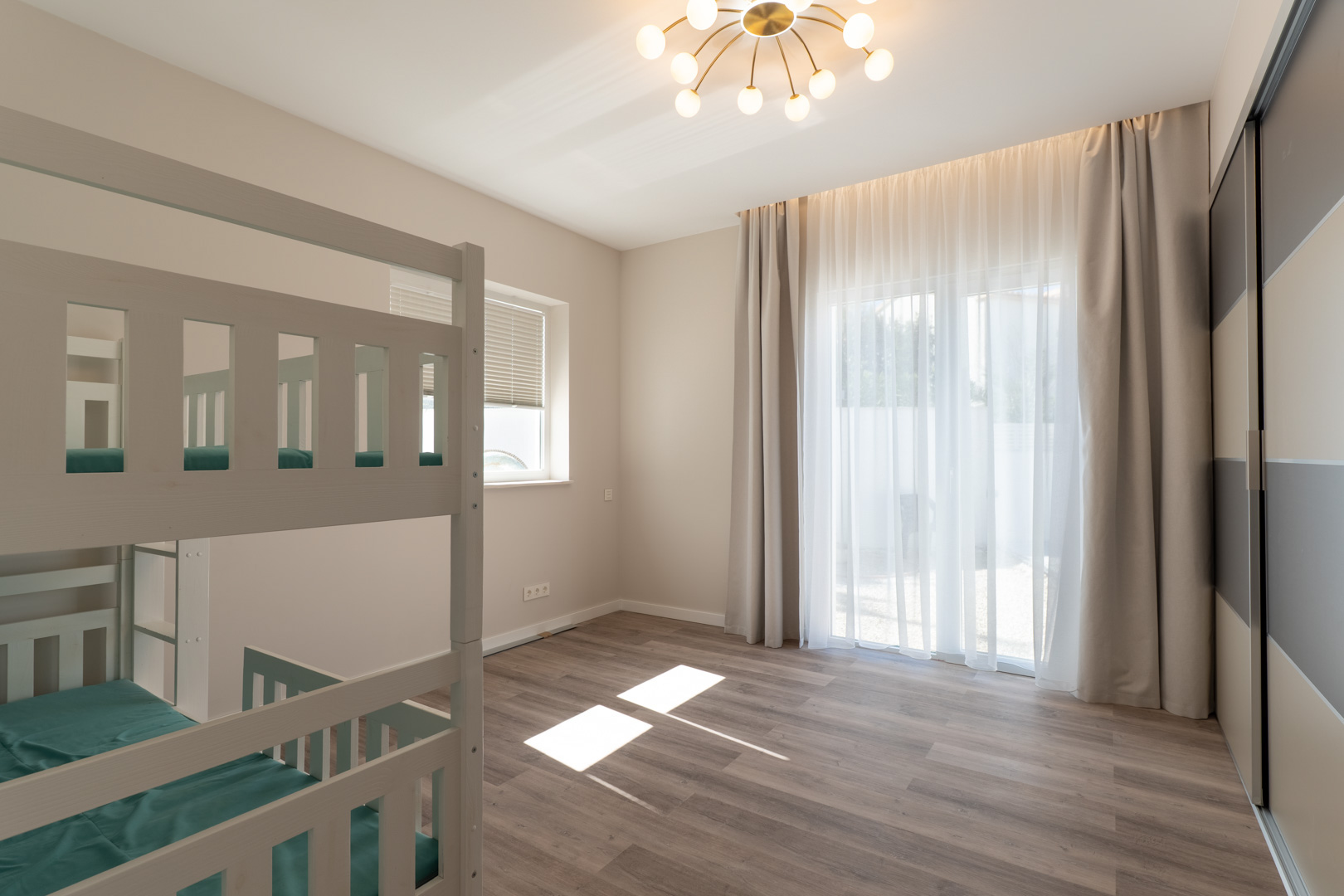 Schlafzimmer mit Etagenbetten aus weißem Holz, Laminatboden und Einbauschrank. Natürliches Licht flutet durch bodentiefe Fenster mit hellen Vorhängen.
