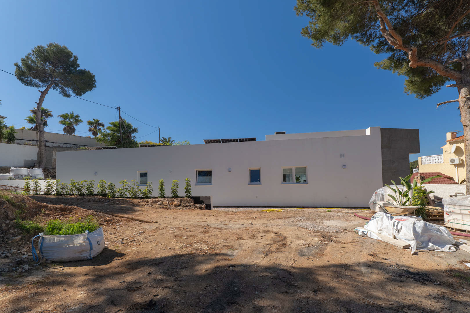 Seitenfassade der minimalistischen Villa mit Solarmodulen und dem Außenbereich in der Vorbereitungsphase, umgeben von altem Pinienbestand.