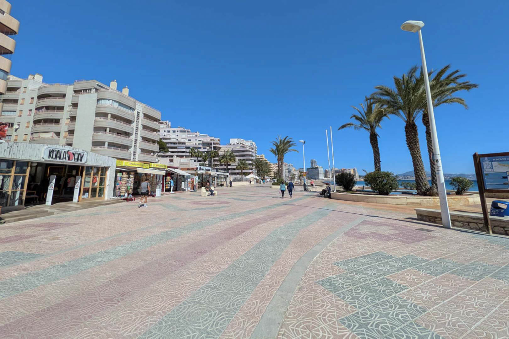 Strandpromenade von Calpe mit gemustertem Steinpflaster, gesäumt von lokalen Geschäften, Palmen und Wohngebäuden bei klarem Himmel.
