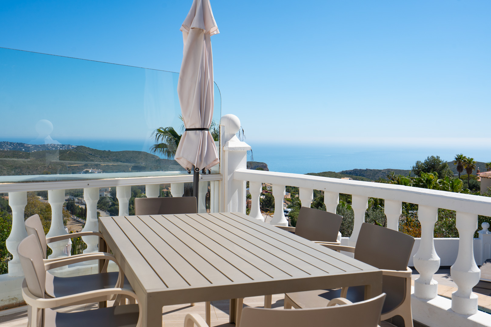 Außen-Essbereich auf der Terrasse mit Lamellentisch und Stühlen, weißer Balustrade und Glaspaneelen mit Panoramablick auf das Mittelmeer und die Küste.