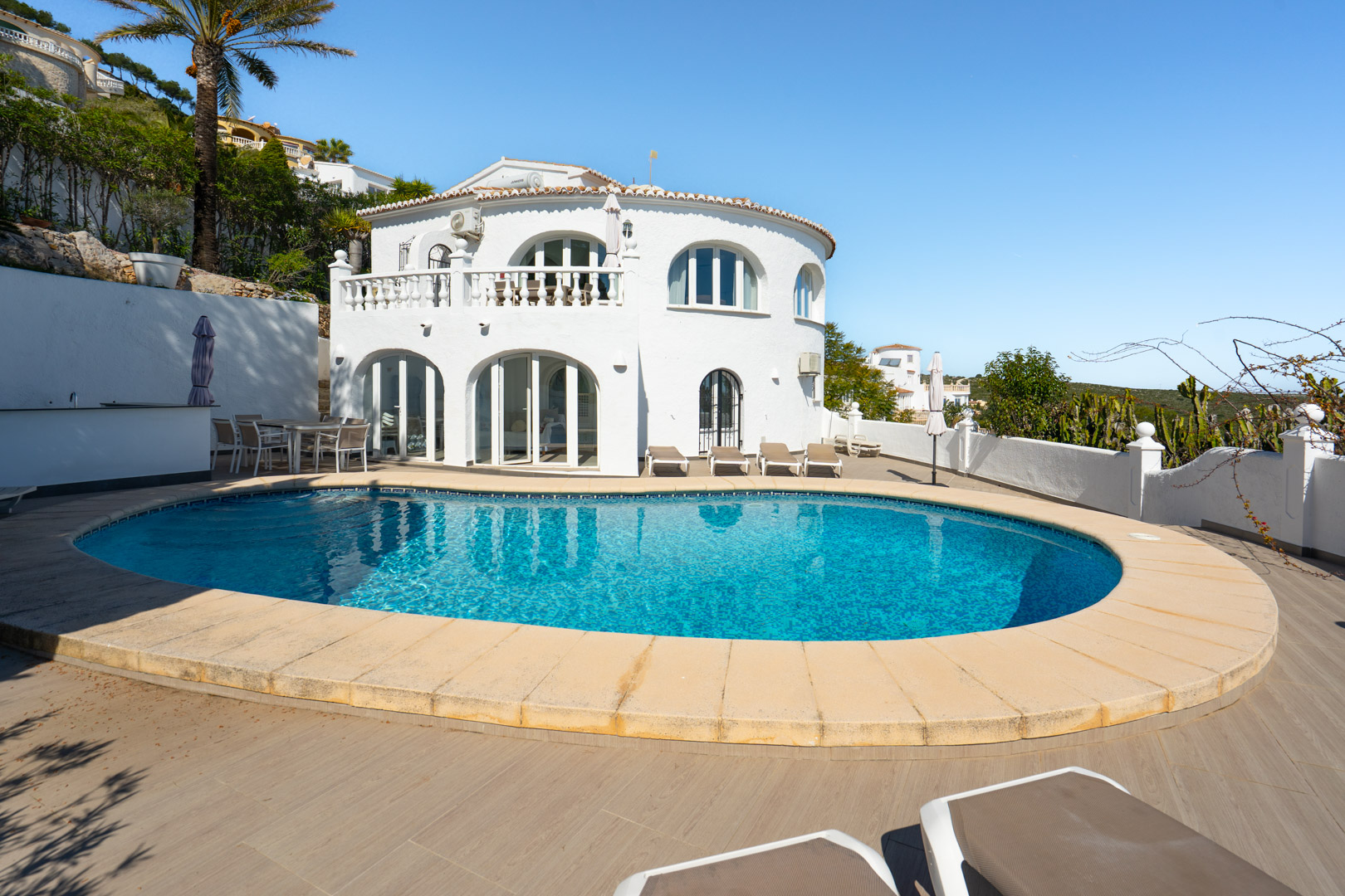 Außenansicht der Villa mit geschwungenem Pool, großzügiger Terrasse in Holzoptik und weißer Fassade mit Rundbogenfenstern und traditioneller Balustrade.
