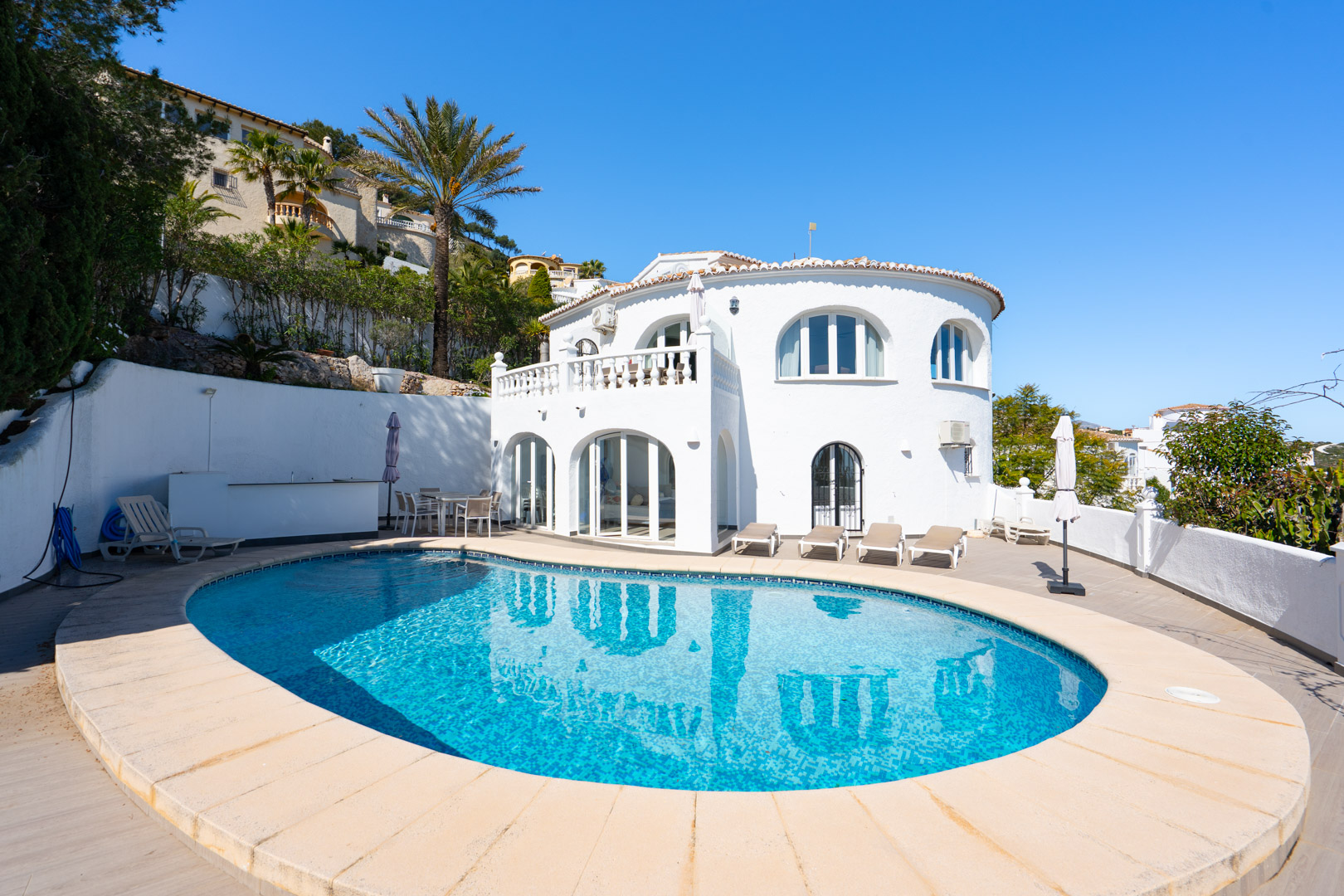Außenansicht der Villa mit ovalem Pool, großzügiger Sonnenterrasse und traditioneller weißer Fassade mit Rundbögen und verglaster Naya.
