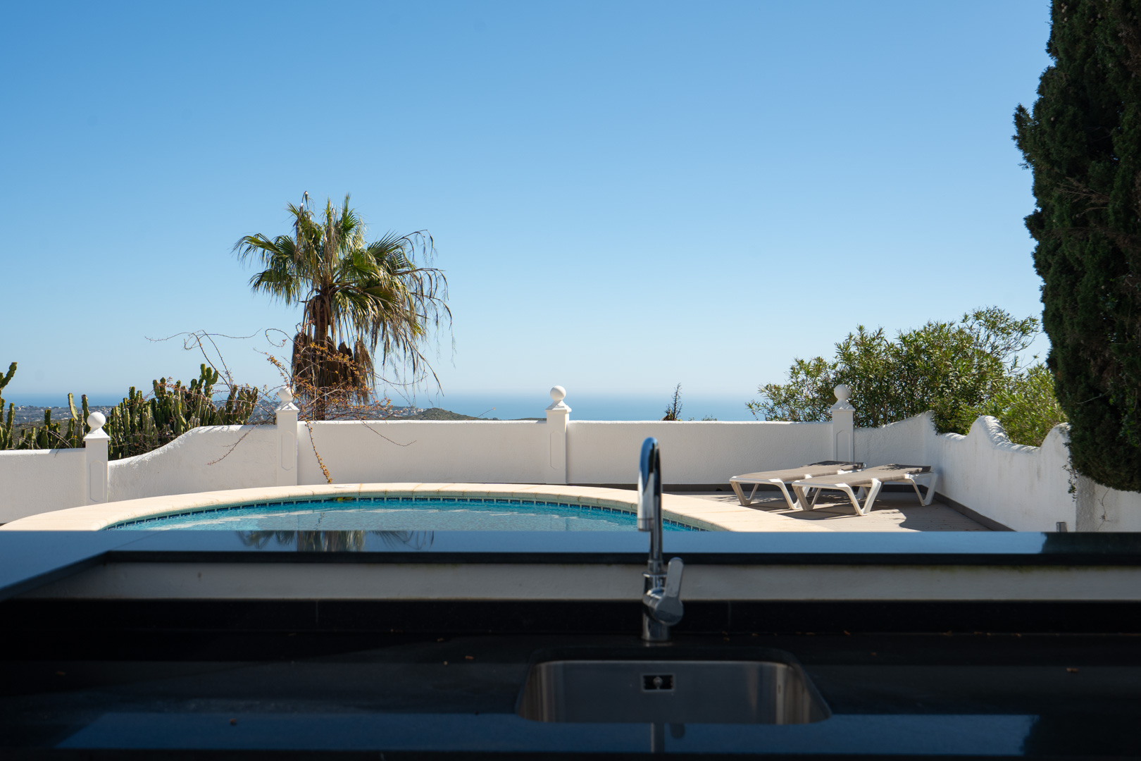 Außenküche mit dunkler Arbeitsplatte und Spüle, mit Blick auf den geschwungenen Pool und die Terrasse mit Meerfernsicht.