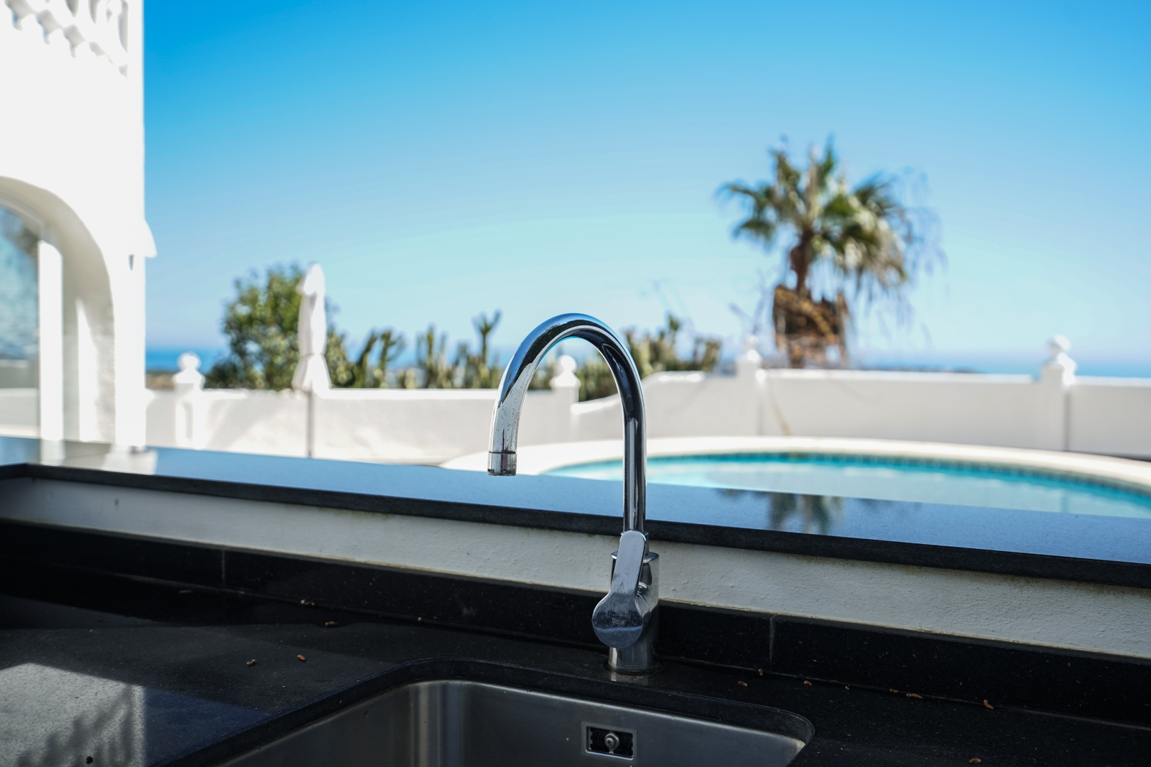 Außenküche mit schwarzer Granitarbeitsplatte und Edelstahlspüle, mit direktem Blick auf den privaten Pool und die Palmen.
