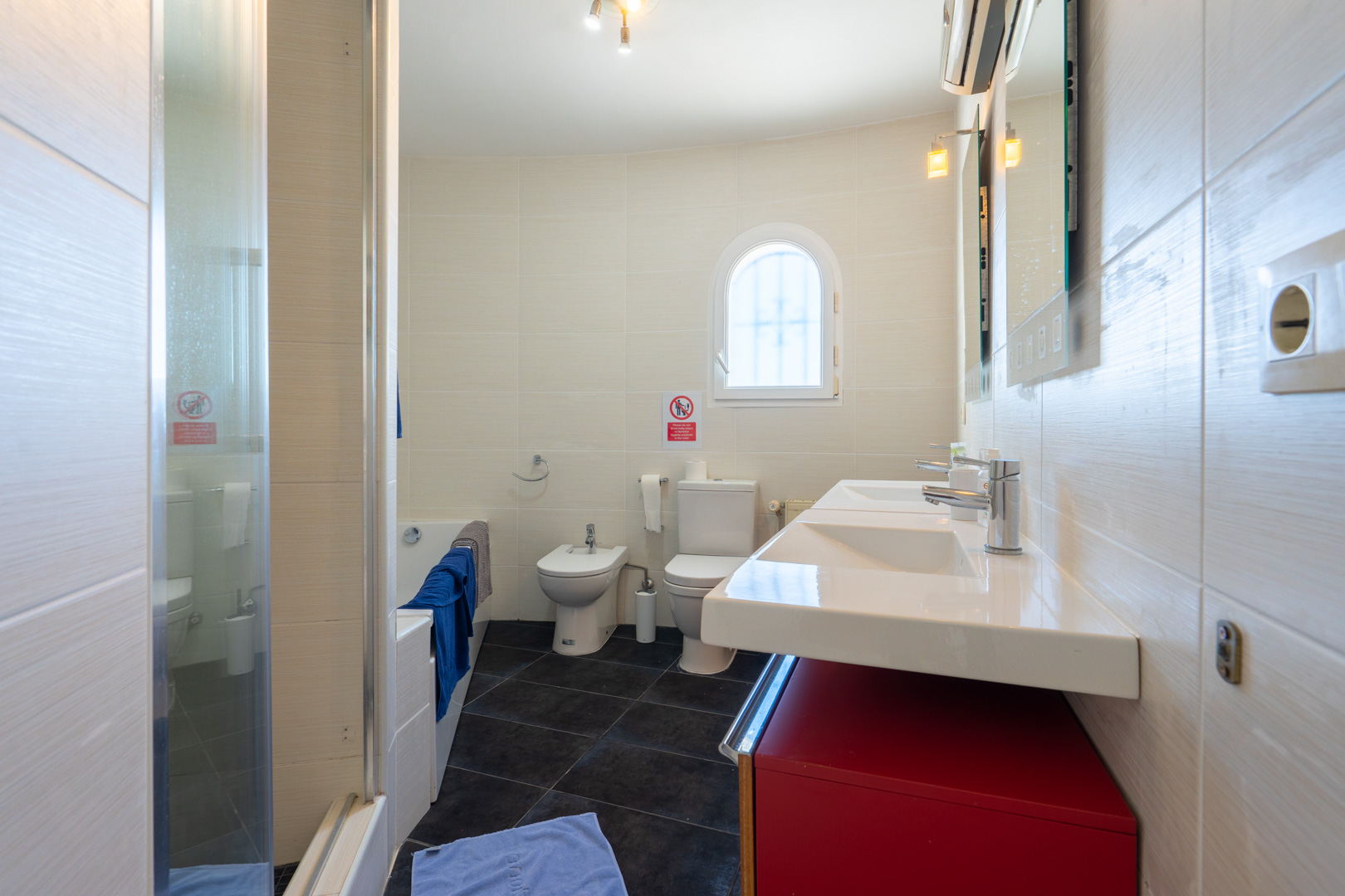 Badezimmer mit Doppelwaschbecken auf rotem Unterschrank, Bidet, WC und Badewanne. Helle Wandfliesen und dunkler Boden ergänzen das Rundbogenfenster.