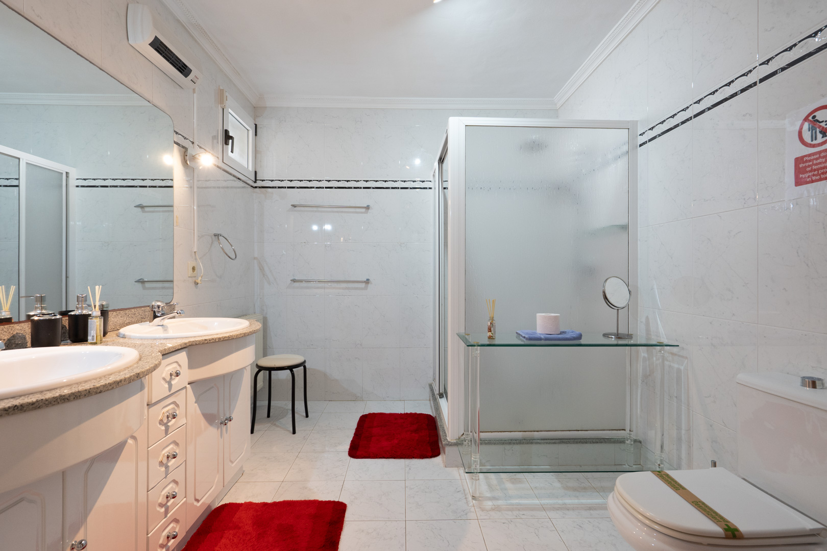 Badezimmer mit Doppelwaschtisch auf Granitplatte, großem Spiegel und Dusche mit Milchglaskabine. Ausgestattet mit Split-Klimaanlage und weißen Fliesenwänden.