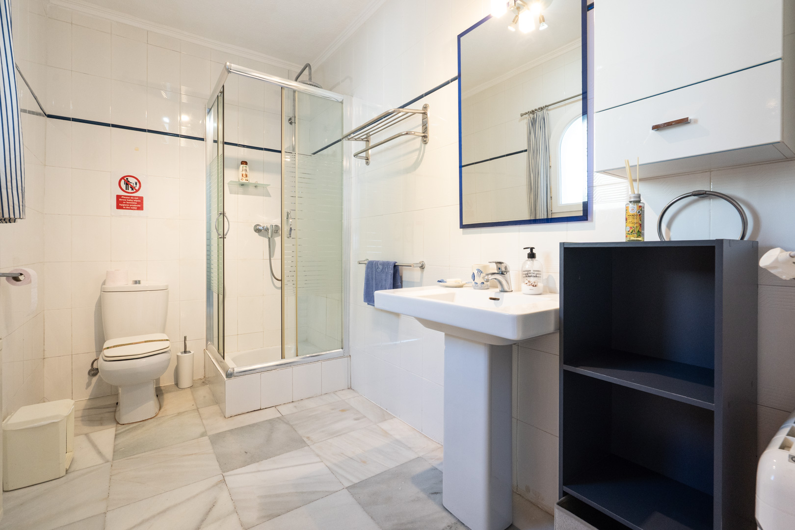 Badezimmer mit weißen Wandfliesen, Marmorboden, einer Eckduschkabine aus Glas und einem Säulenwaschbecken mit blau gerahmtem Spiegel.