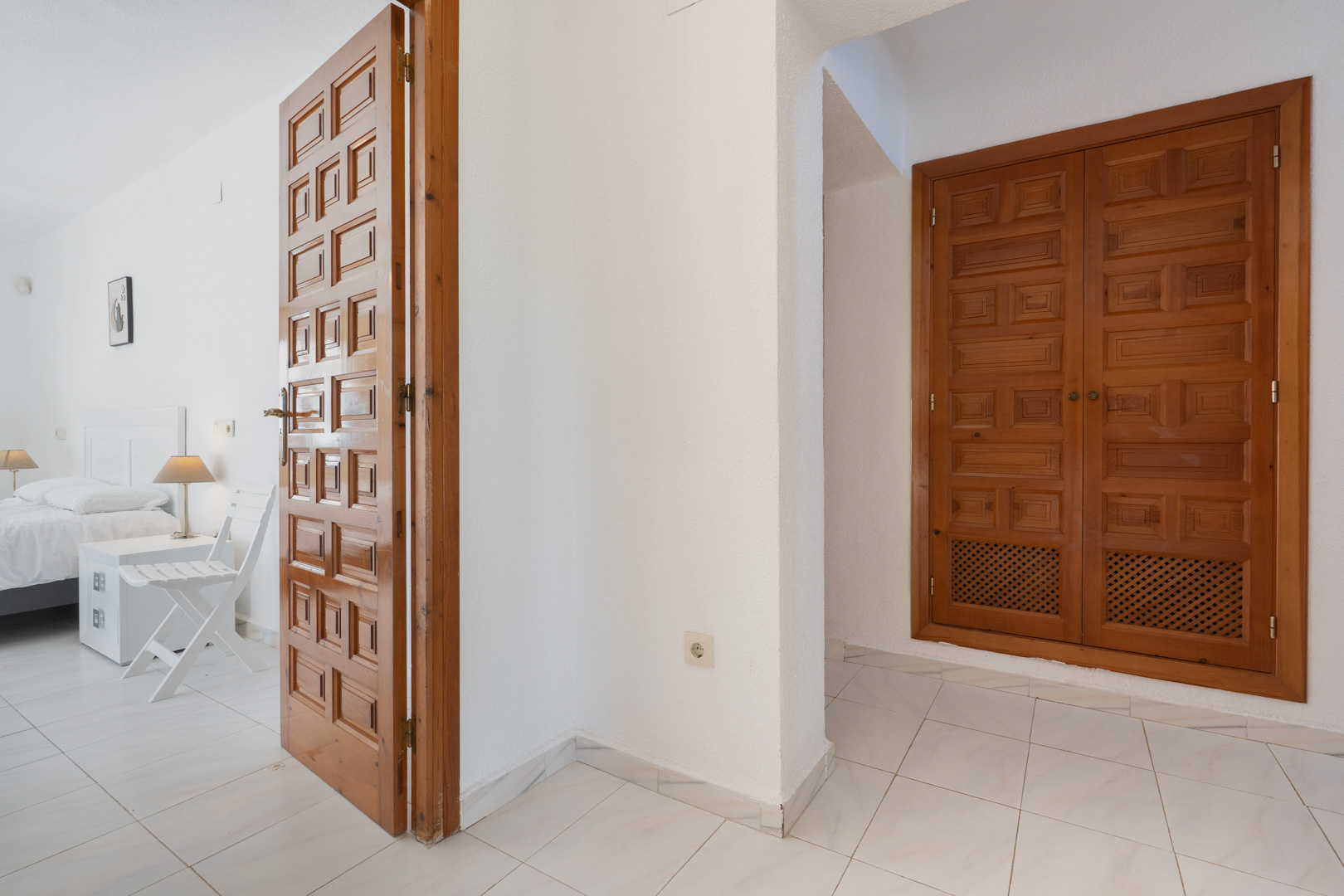 Flur mit traditionell geschnitzten Holztüren und Einbauschränken, der in ein helles Schlafzimmer mit weißen Fliesenböden und schlichtem Design führt.
