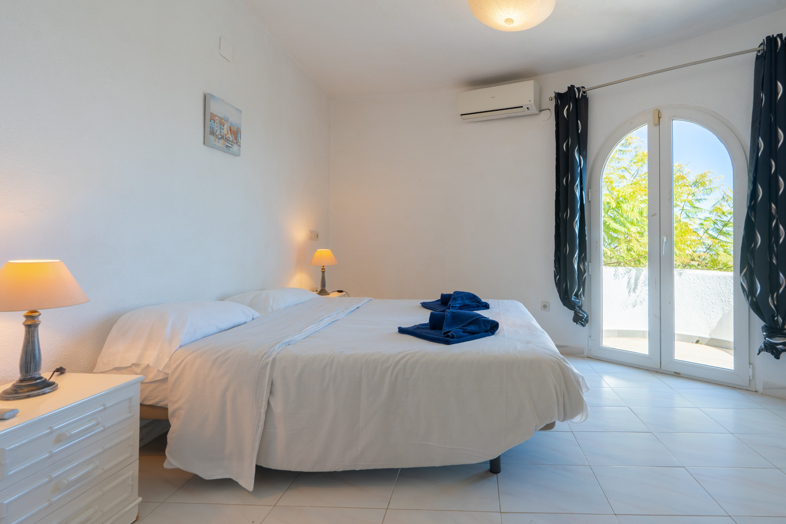 Helles Schlafzimmer mit Doppelbett, Klimaanlage und einer bogenförmigen Glastür, die direkt auf die Terrasse führt.