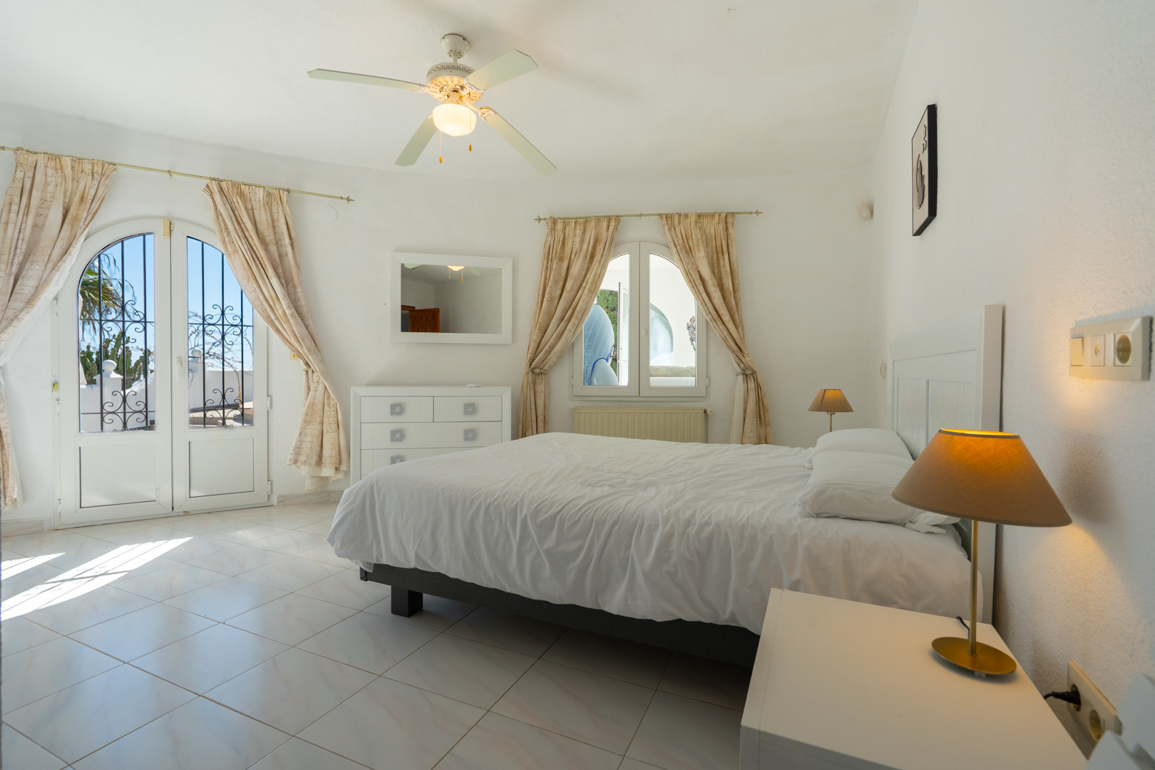 Helles Schlafzimmer mit Fliesenboden, Deckenventilator und direktem Außenzugang durch eine bogenförmige Glastür mit dekorativem Sicherheitsgitter.