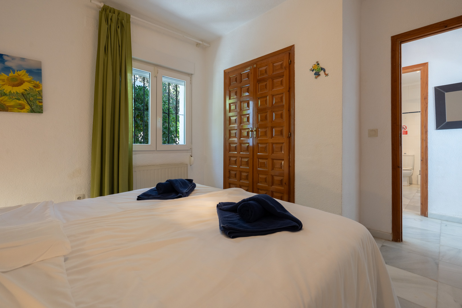 Helles Schlafzimmer mit geschnitztem Einbauschrank aus Holz, grünen Vorhängen und Zugang zu einem Badezimmer mit Marmorfliesen.