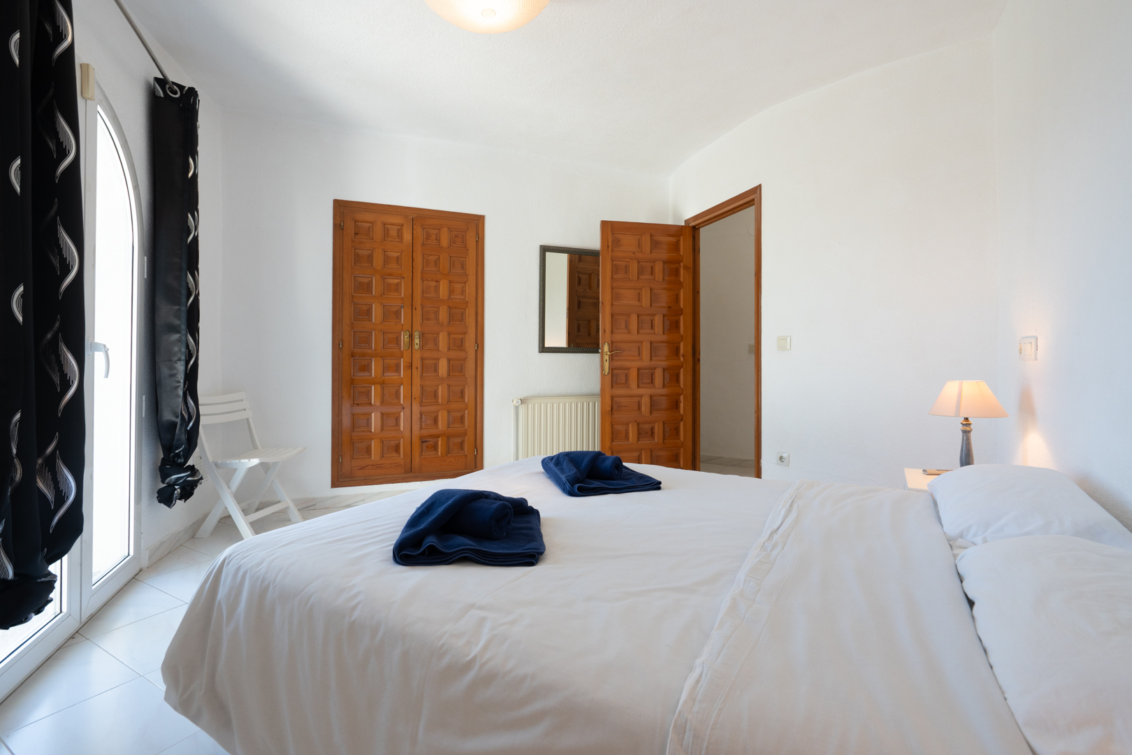 Helles Schlafzimmer mit traditionellen Einbauschränken aus Holz, einer bogenförmigen Glastür zum Außenbereich und hellen Fliesenböden.