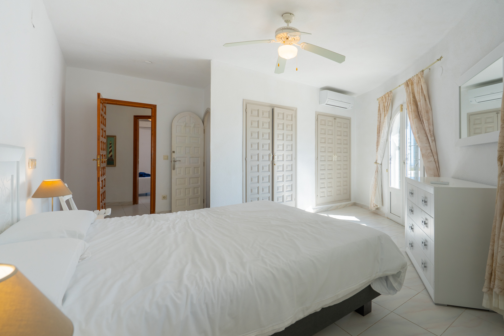 Helles Schlafzimmer mit traditionellen Einbauschränken, Klimaanlage, Deckenventilator und direktem Zugang nach draußen durch eine verglaste Rundbogentür.