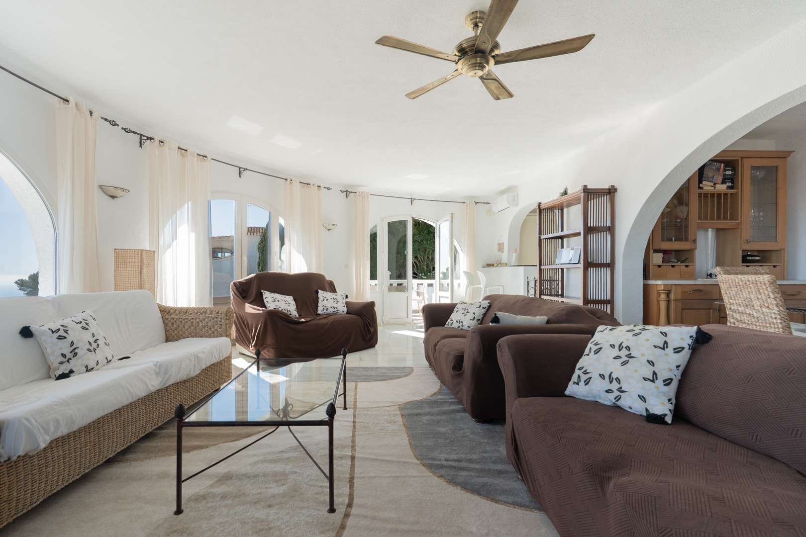 Helles Wohnzimmer mit geschwungener Wand, Rundbogenfenstern und Terrassenzugang. Ausgestattet mit Holzdeckenventilator, Fliesenboden und offenem Rundbogen zur Küche.