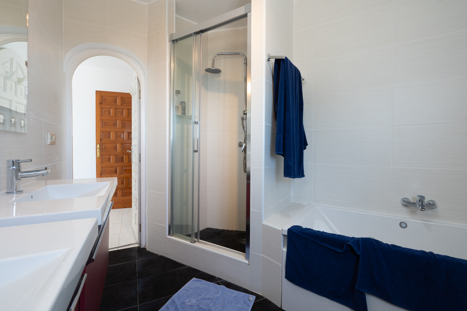 Modernes Badezimmer mit Doppelwaschbecken, Badewanne und separater Dusche mit Glaskabine. Helle Wandfliesen treffen auf einen dunklen Fliesenboden und Rundbogen.