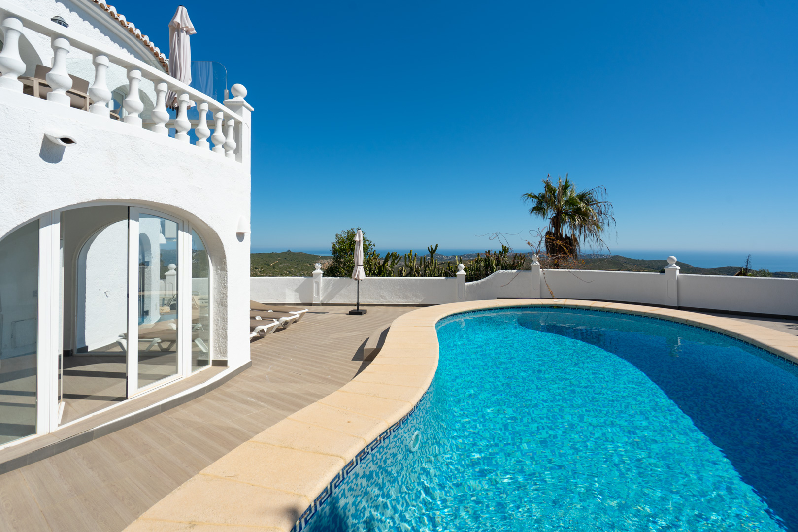 Nierenförmiger Pool mit Mosaikfliesen und großzügiger Sonnenterrasse in Holzoptik, mit Panoramablick auf das Meer und direktem Zugang zur verglasten Naya.