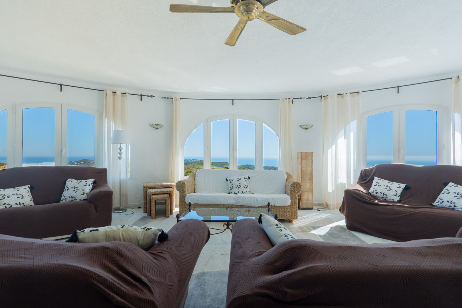 Rundes Wohnzimmer mit bogenförmigen Fenstern und Panoramablick auf das Meer. Ausgestattet mit Deckenventilator, Korbmöbeln und hellen, lichtdurchfluteten Wänden.