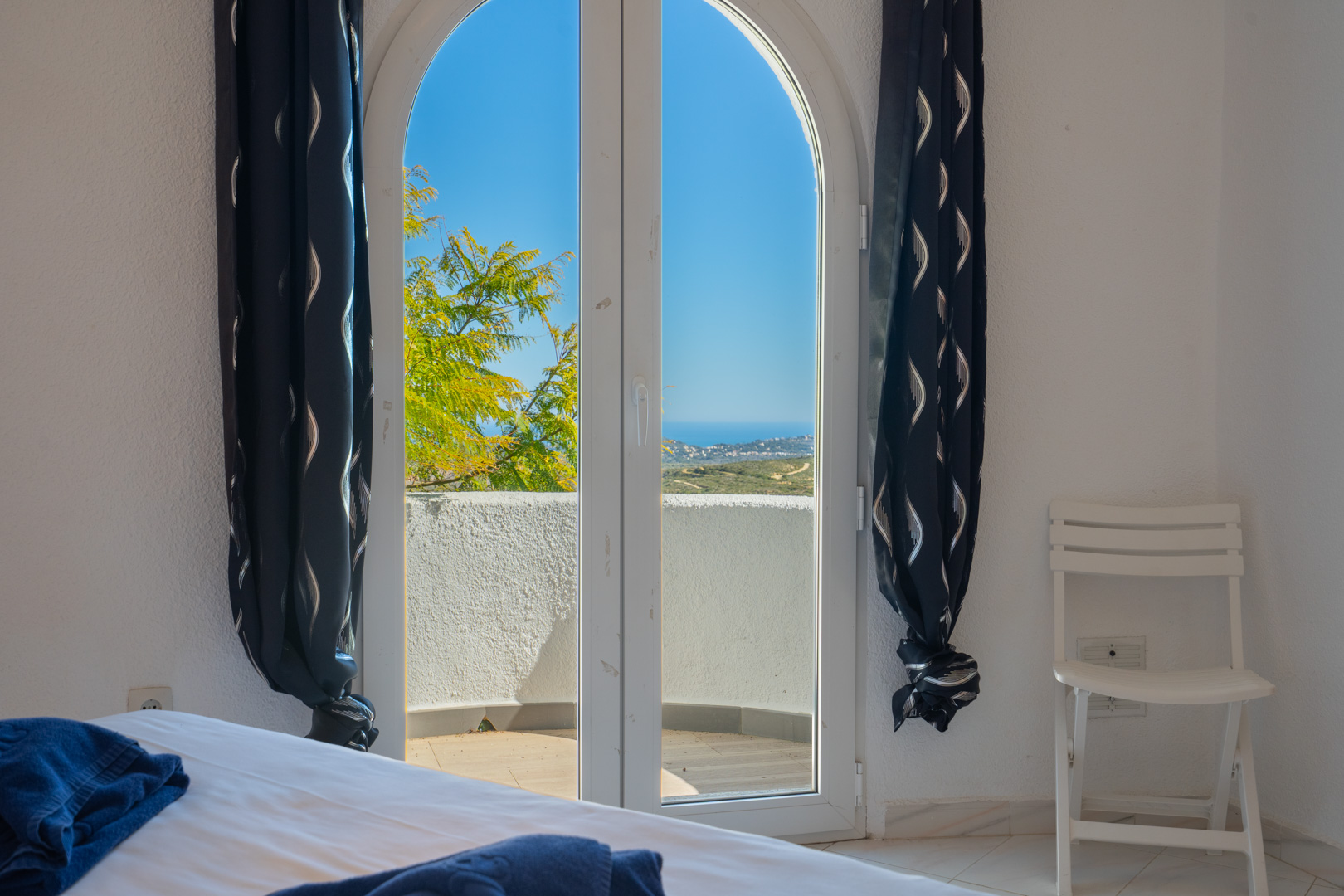 Schlafzimmer mit bogenförmiger Glastür, die auf einen privaten Balkon mit Meerblick führt. Ausgestattet mit weißen Wänden und dunklen Mustervorhängen.