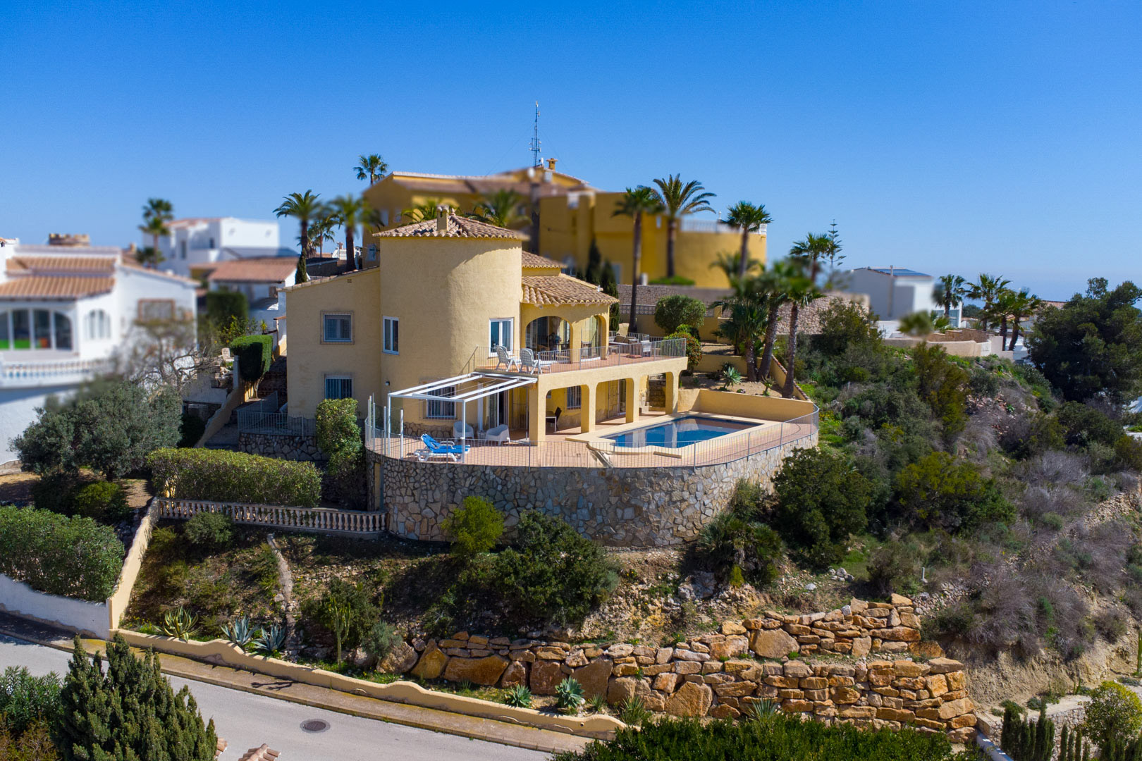 Außenansicht der Villa mit ockerfarbener Fassade, Rundturm und rechteckigem Pool auf einer weitläufigen Terrasse mit Natursteinmauern.