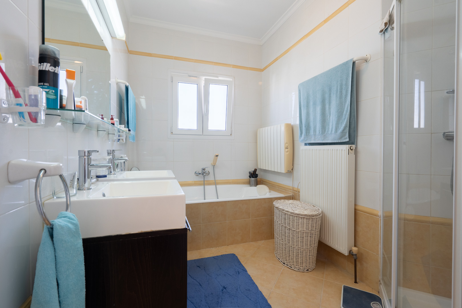 Badezimmer mit Doppelwaschbecken, Einbaubadewanne, separater Dusche und weißen Wandfliesen mit gelber Zierleiste.