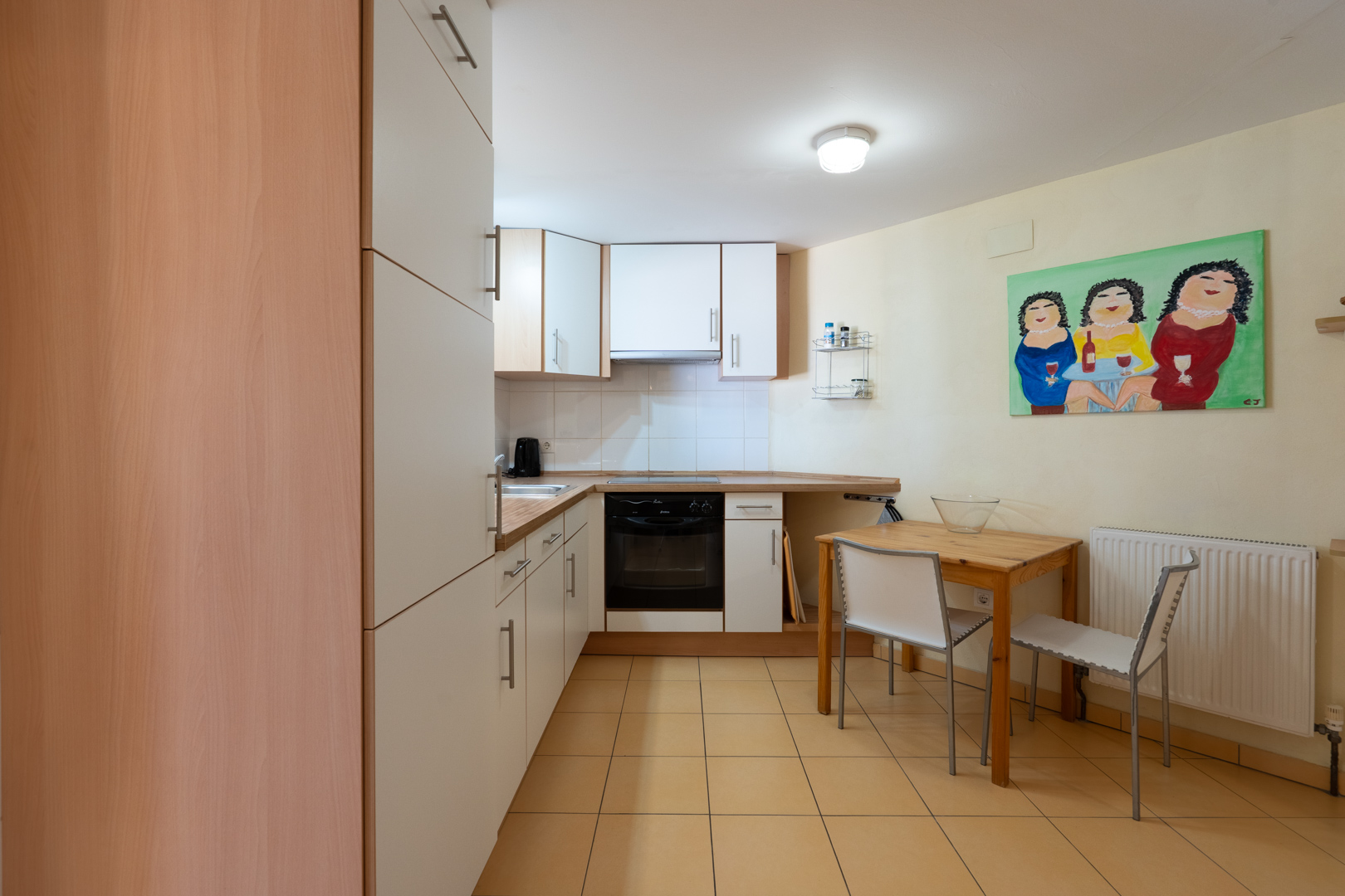 Funktionale Küche der Einliegerwohnung mit weißen Schränken, Arbeitsplatten in Holzoptik, kompaktem Essbereich und beigefarbenen Bodenfliesen.