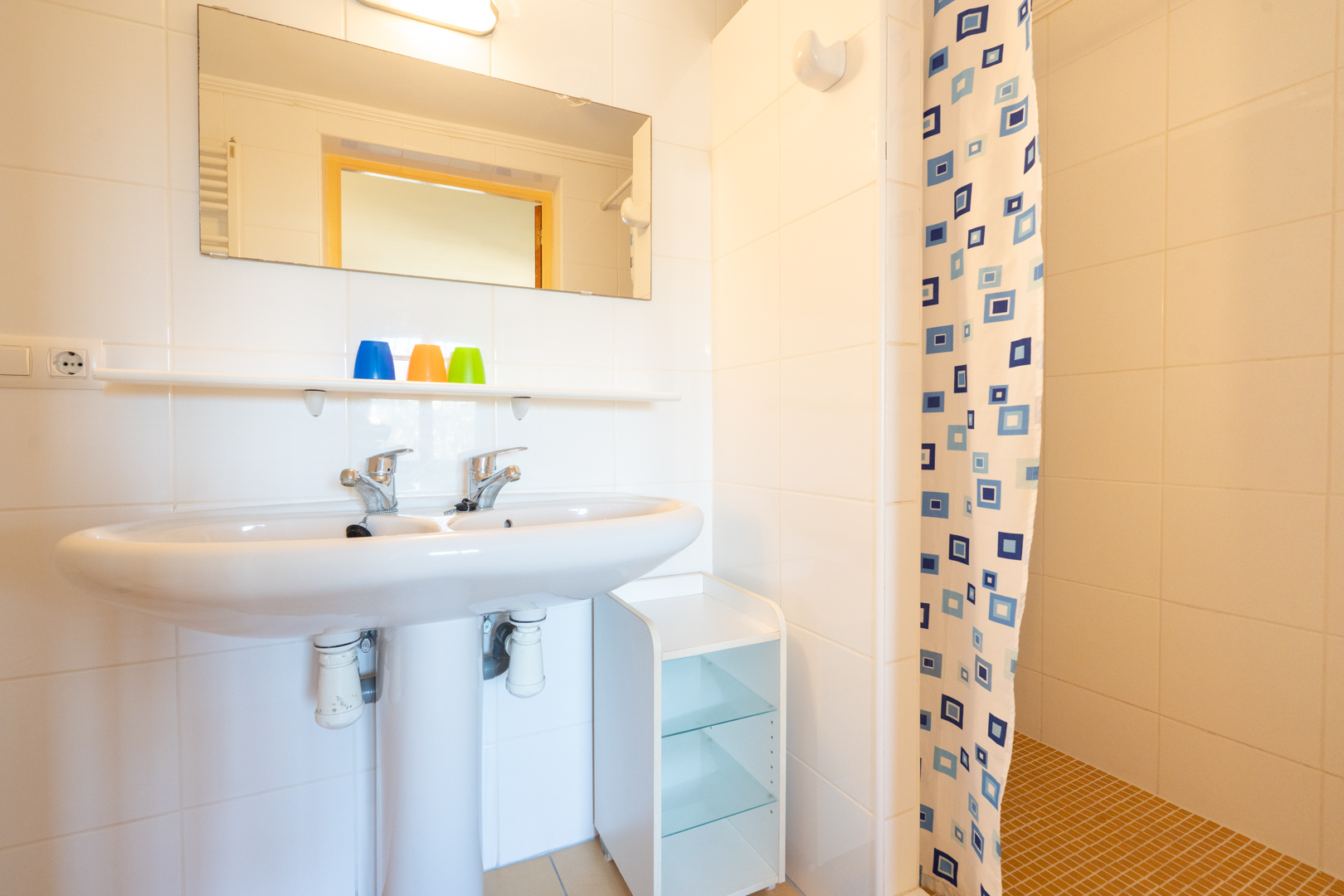 Funktionales Badezimmer mit Doppelwaschbecken auf Säule, weißen Wandfliesen und Duschbereich mit gemustertem Vorhang und ockerfarbenem Fliesenboden.