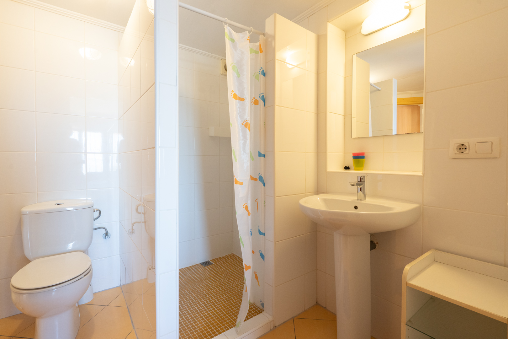 Funktionales Badezimmer mit weißen Wandfliesen, ebenerdiger Dusche mit Vorhang, Standwaschbecken und klassischen Sanitärobjekten.