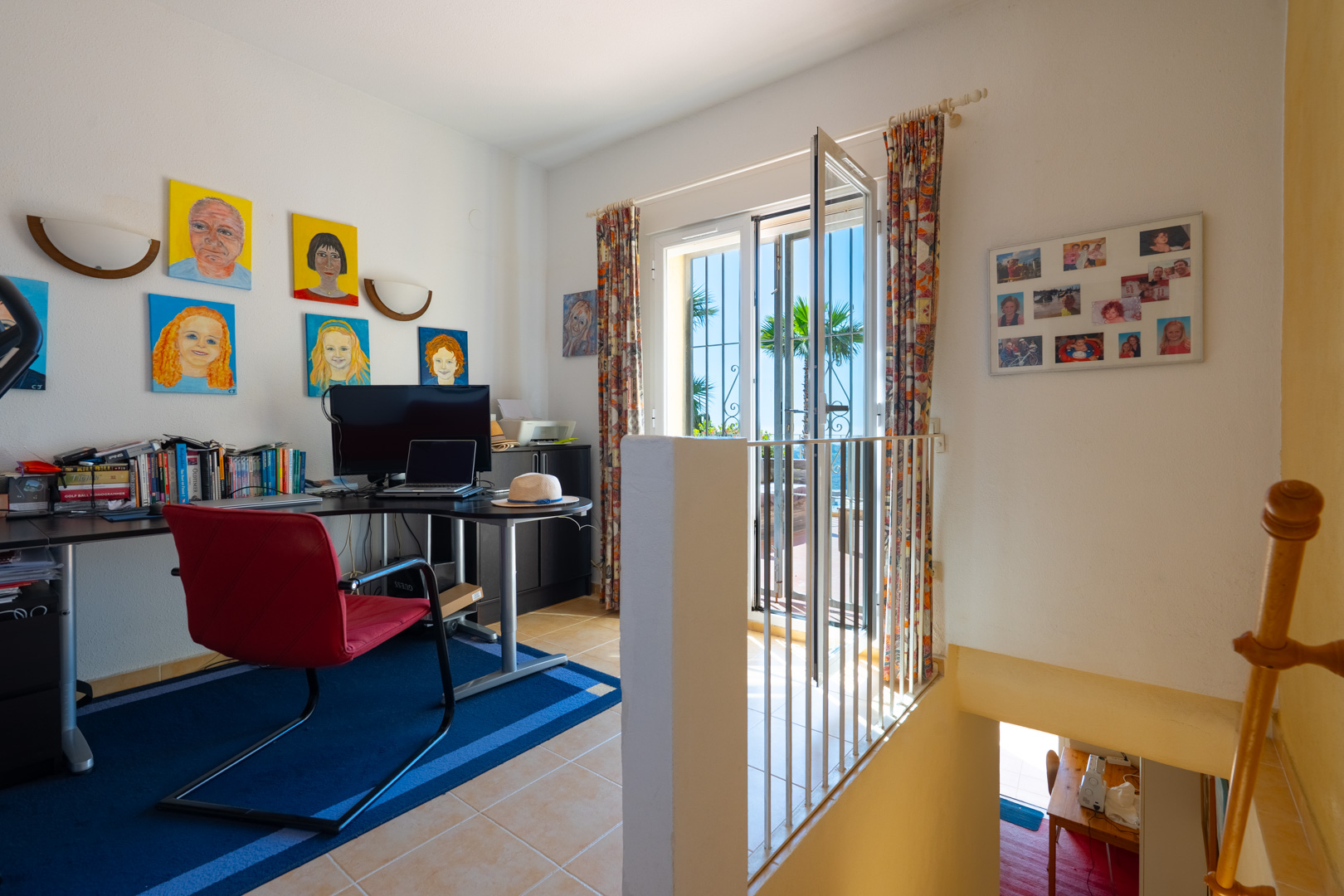 Hell eingerichtetes Arbeitszimmer mit Eckschreibtisch, rotem Stuhl und Terrassenzugang. Inklusive Treppenaufgang zum Untergeschoss und Fliesenboden.