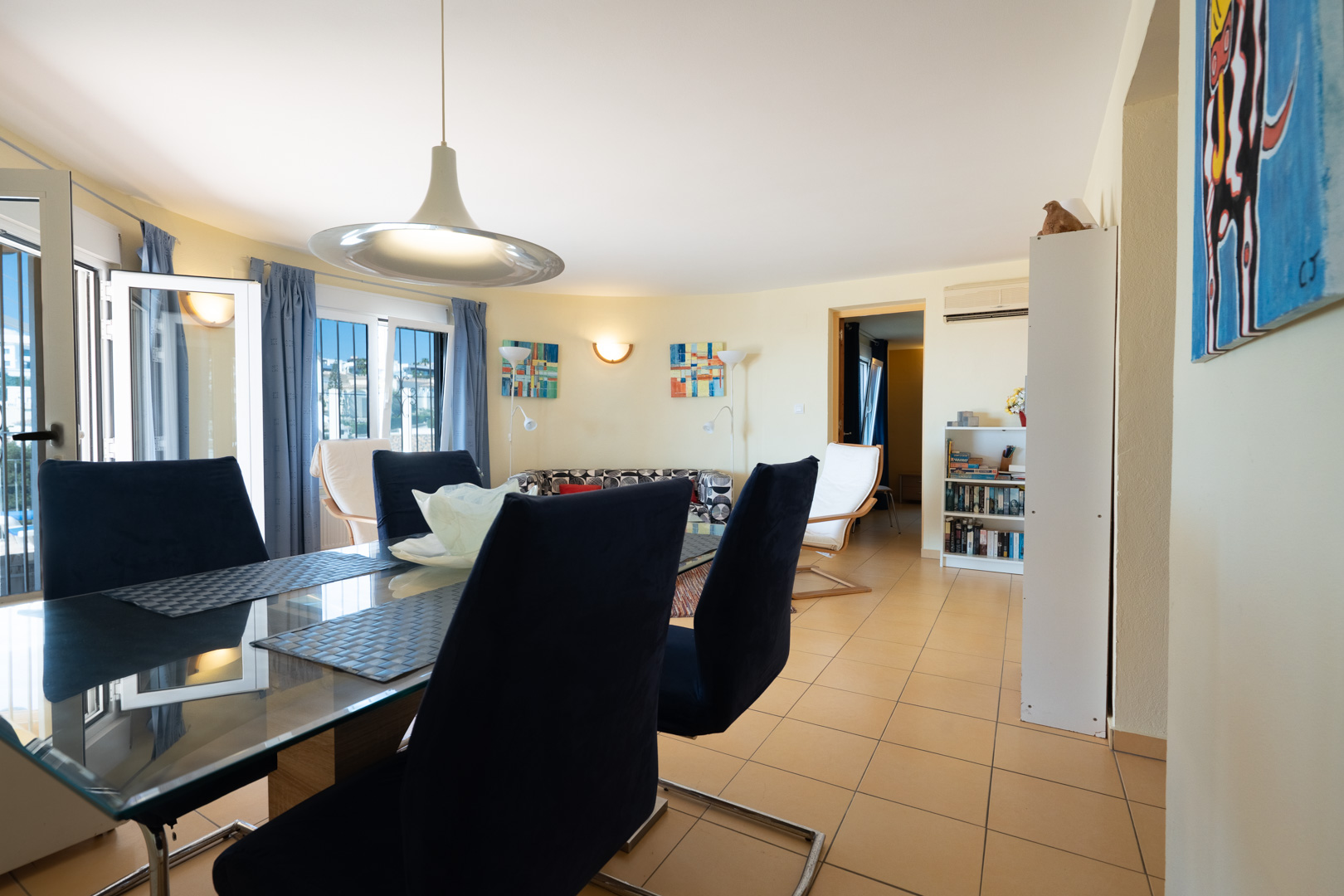 Heller Essbereich mit Glastisch und blauen Samtstühlen, integriert in ein offenes Wohnzimmer mit Fliesenboden und direktem Zugang zum Außenbereich.