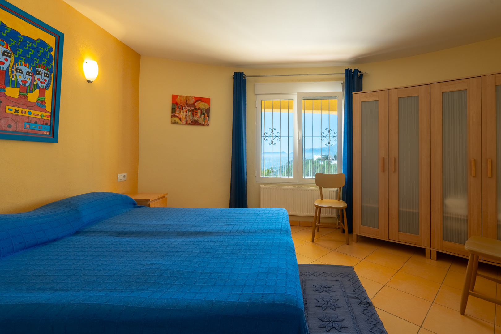 Helles Schlafzimmer mit Doppelbett, ockerfarbenen Wänden, Holzschrank mit Milchglaseinsätzen und Fenster mit Küstenblick.
