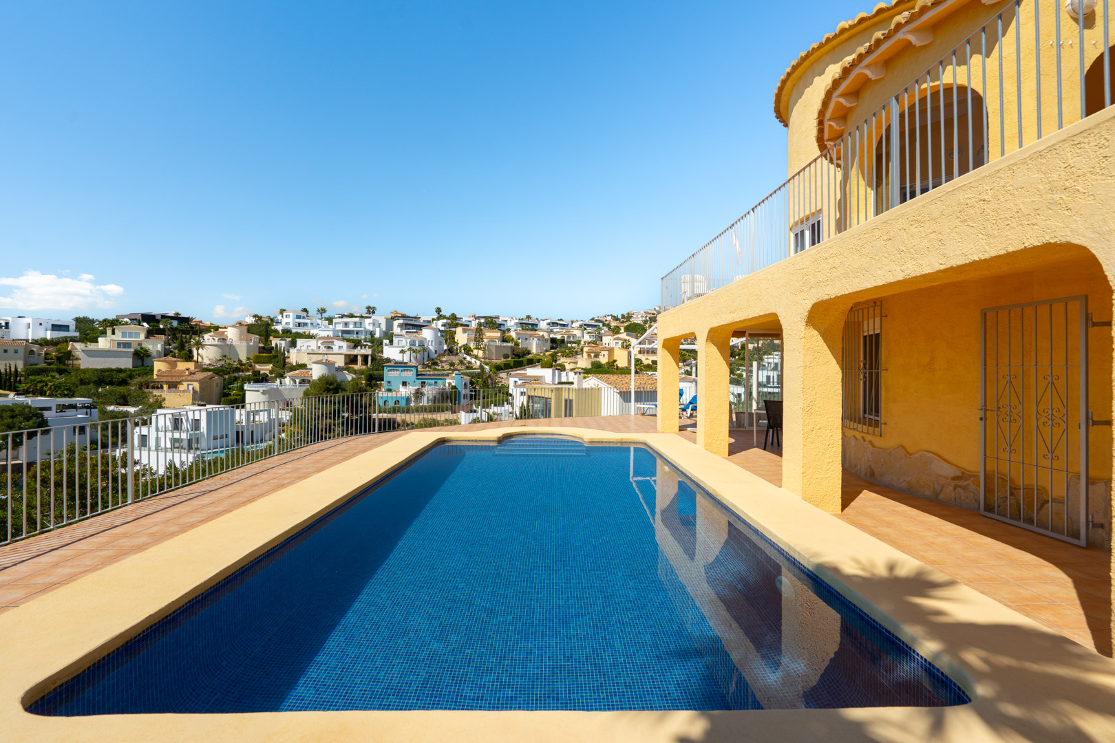 Rechteckiger Swimmingpool mit blauen Mosaikfliesen, umgeben von einer großzügigen Fliesenterrasse und einer gelben Fassade mit Rundbögen.