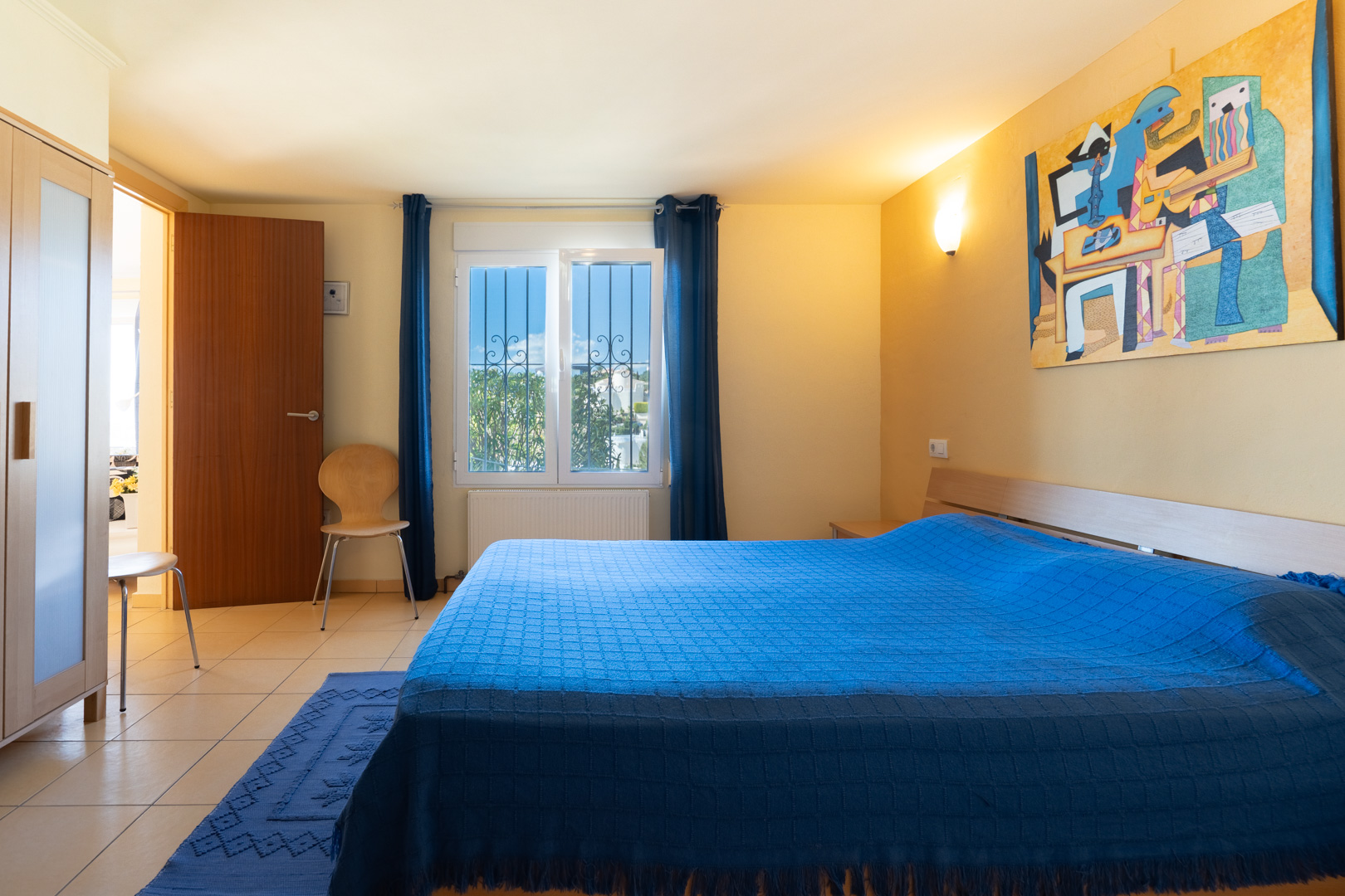 Schlafzimmer mit Doppelbett, blauen Textilien, Holzschrank und Fliesenboden. Ein Fenster mit Sicherheitsgittern lässt natürliches Licht herein.