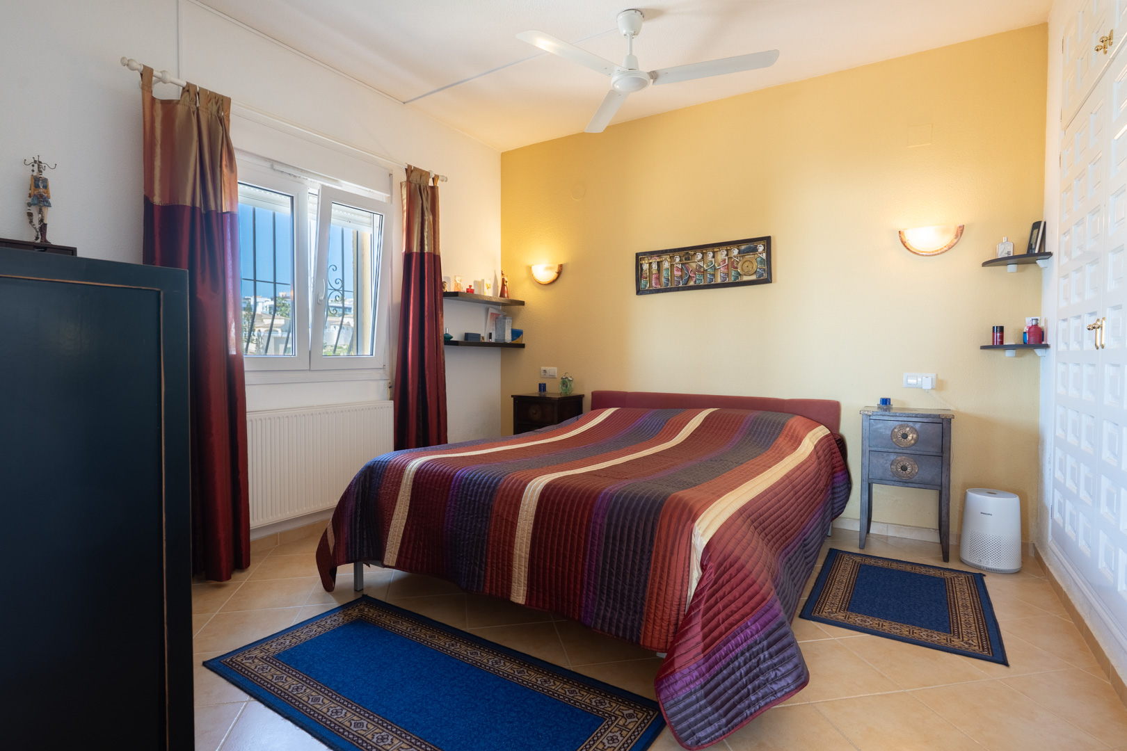 Schlafzimmer mit Doppelbett, gelber Akzentwand, Deckenventilator und weißen Einbauschränken. Ausgestattet mit Heizkörper und Keramikfliesenboden.
