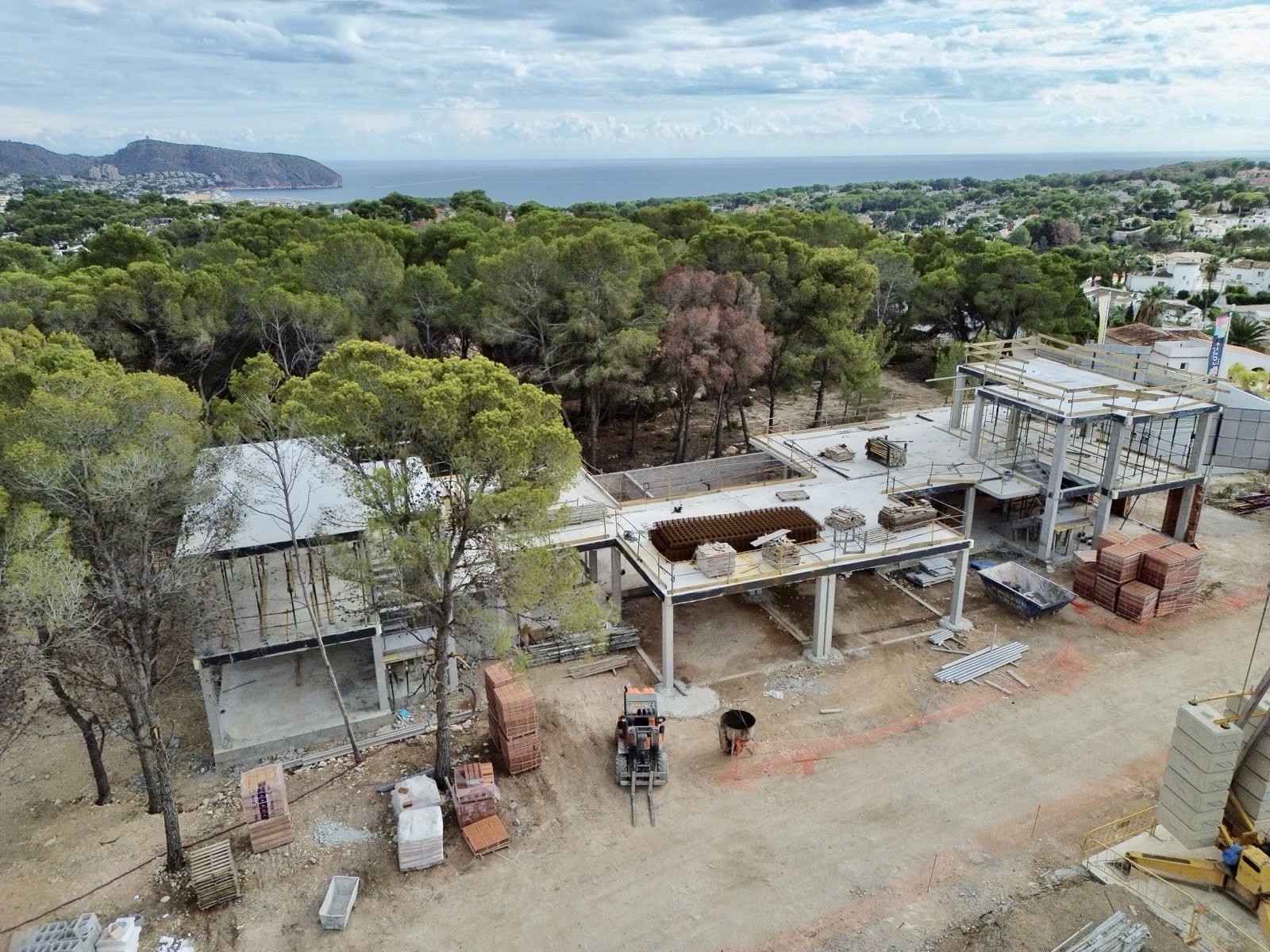 Luftaufnahme der im Bau befindlichen Villa, die ihre Betonstruktur und Baumaterialien zeigt. Im Hintergrund sind das Mittelmeer und die bewaldete Landschaft zu sehen.