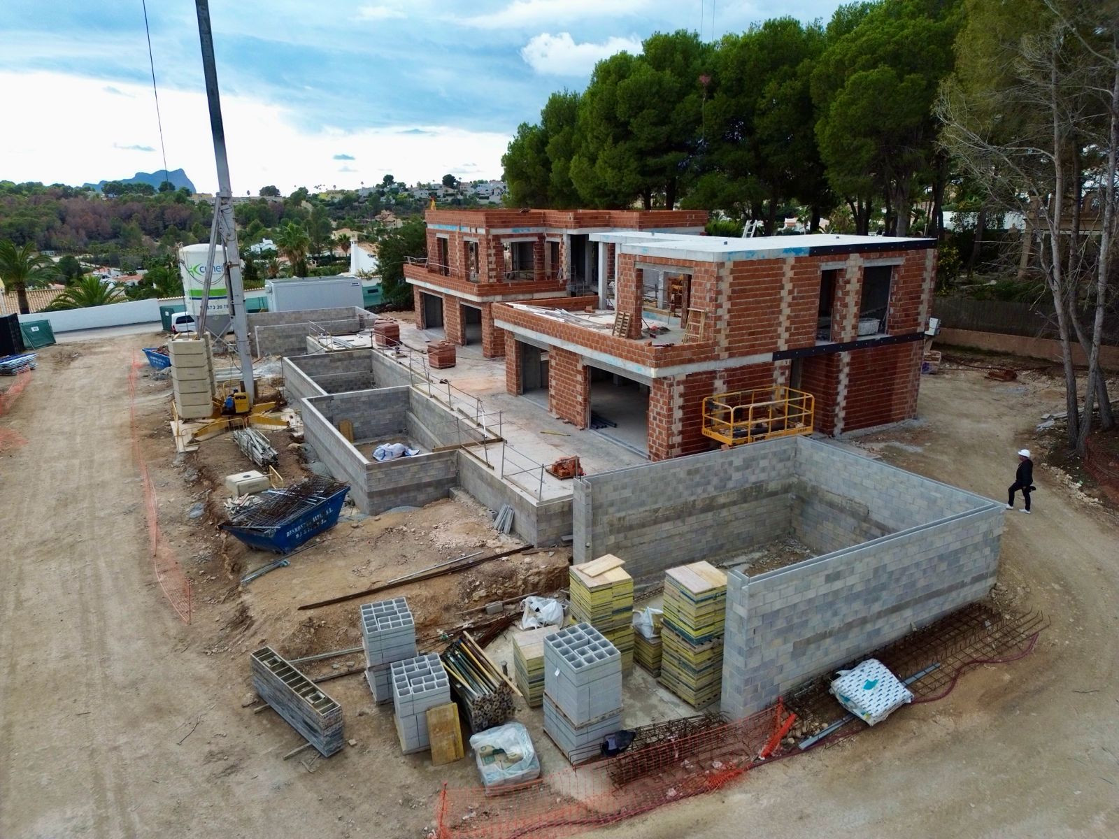 Luftaufnahme der Villenbaustelle, mit mehreren im Bau befindlichen Ziegelstrukturen, ausgehobenen Betonfundamenten und Baumaterialien. Ein Kran ist sichtbar, mit fernen grünen Hügeln.