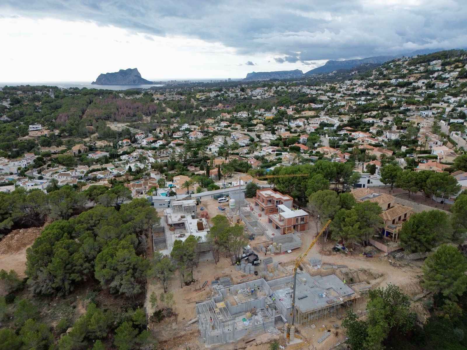 Luftaufnahme von Villen im Bau in einem Wohngebiet von Moraira, mit Kränen und Fundamenten, umrahmt vom Meer und dem Peñón de Ifach in der Ferne.