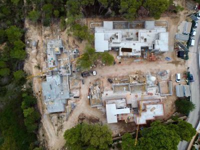 Luftaufnahme eines Villenkomplexes im Bau, mit Fundamenten, Betonplatten und zwei Kränen. Baumaterialien und Fahrzeuge sind auf dem Gelände sichtbar.