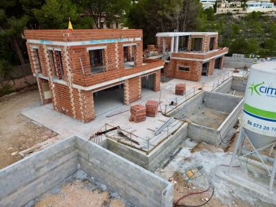 Luftaufnahme eines Villenprojekts im Bau, mit dem Mauerwerk zweier Hauptstrukturen und Betonblockfundamenten, eingebettet in eine grüne Hügellandschaft.