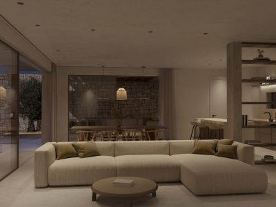 Offener Wohn- und Essbereich mit modularem Sofa, rundem Couchtisch und Zugang zu einer Steinterrasse mit Olivenbaum. Integrierte Küche im Hintergrund erkennbar.