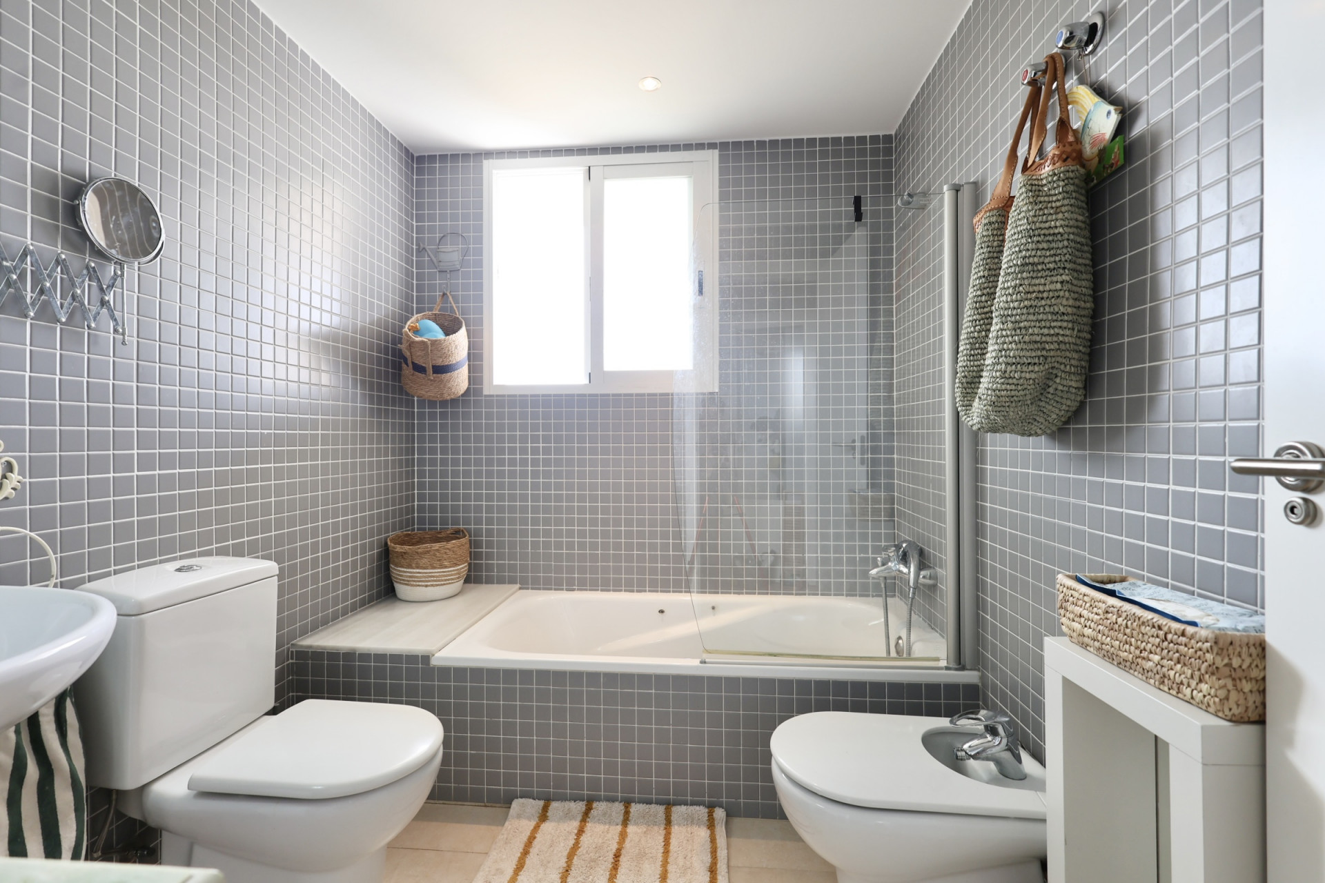 Badezimmer mit grauen Fliesenwänden, Badewanne mit Duschwand und dekorativen Elementen aus Korbweide.