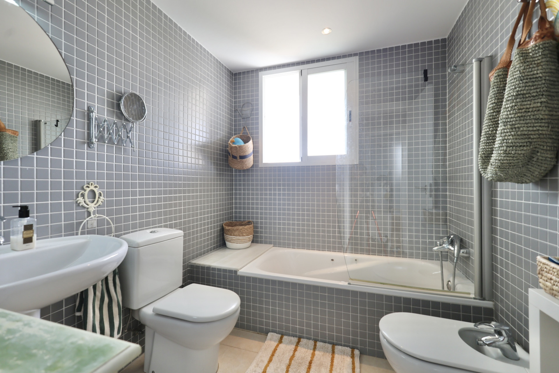 Badezimmer mit grauen quadratischen Fliesen, Waschtisch, Toilette und Bidet. Unter einem Fenster befindet sich eine weiße Badewanne.