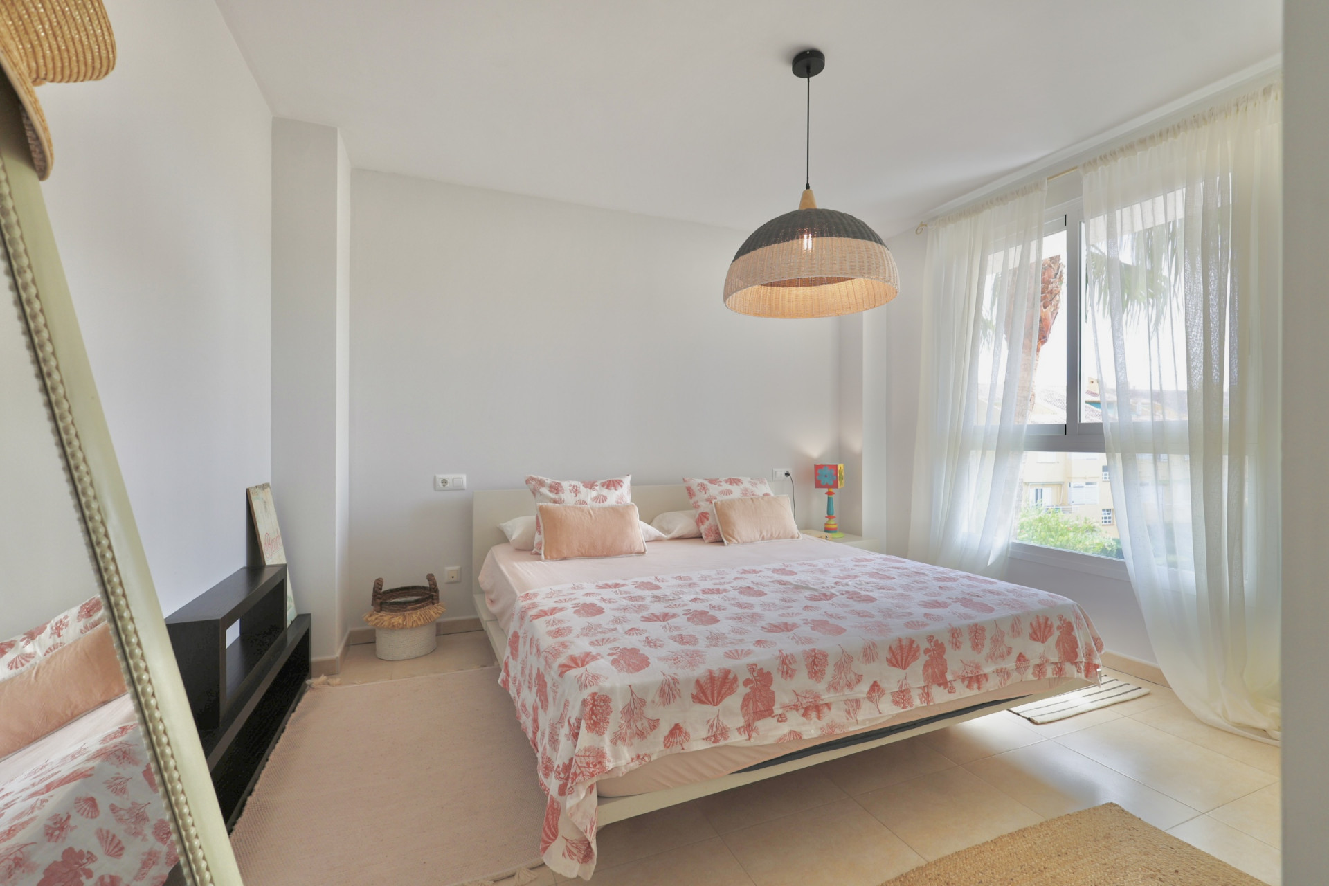 Helles Schlafzimmer mit Doppelbett, gemusterter Bettwäsche und einer Hängelampe aus Korbgeflecht.