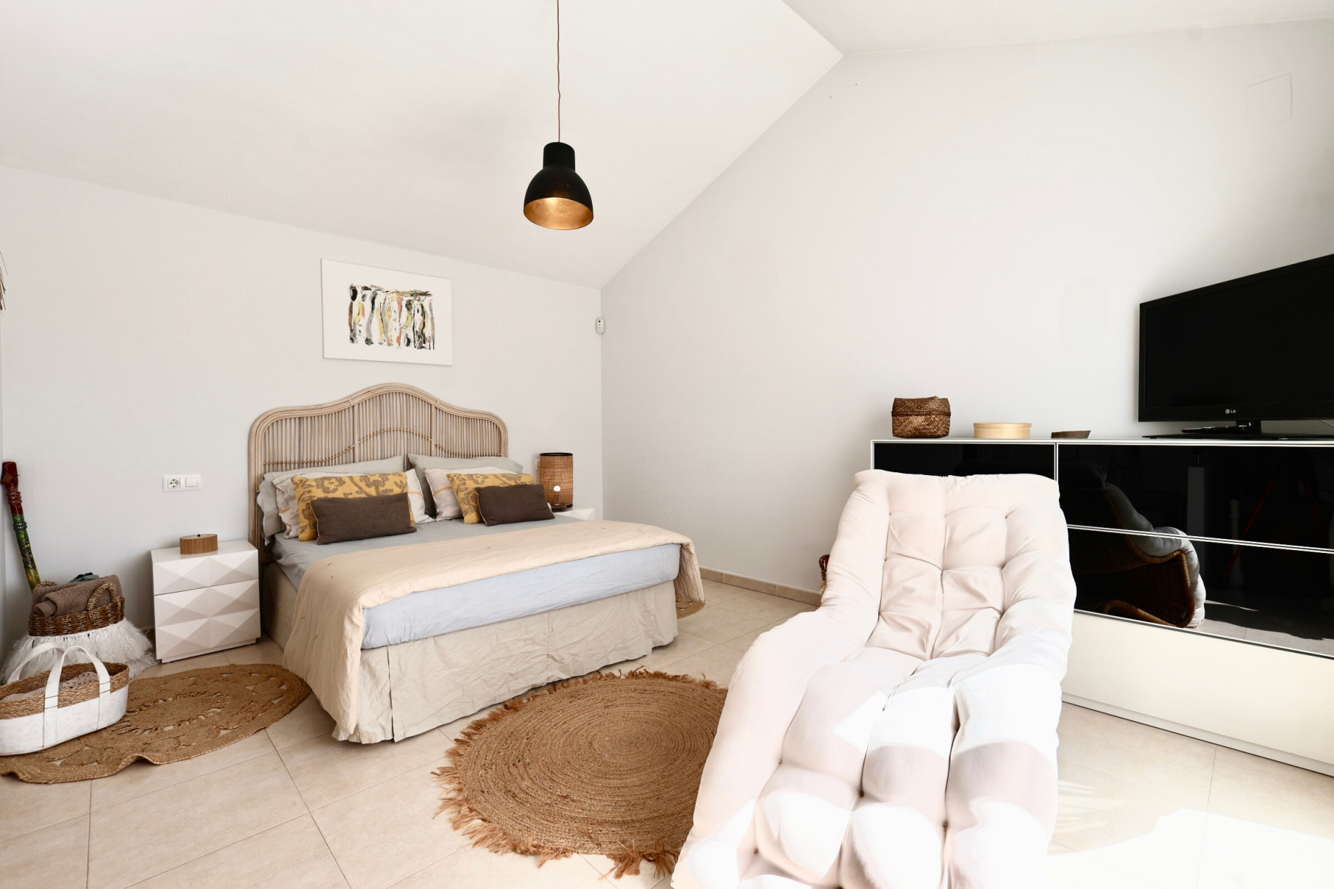 Helles Schlafzimmer mit Rattanbett, Jute-Teppich und Sessel neben einem schwarzen TV-Schrank.