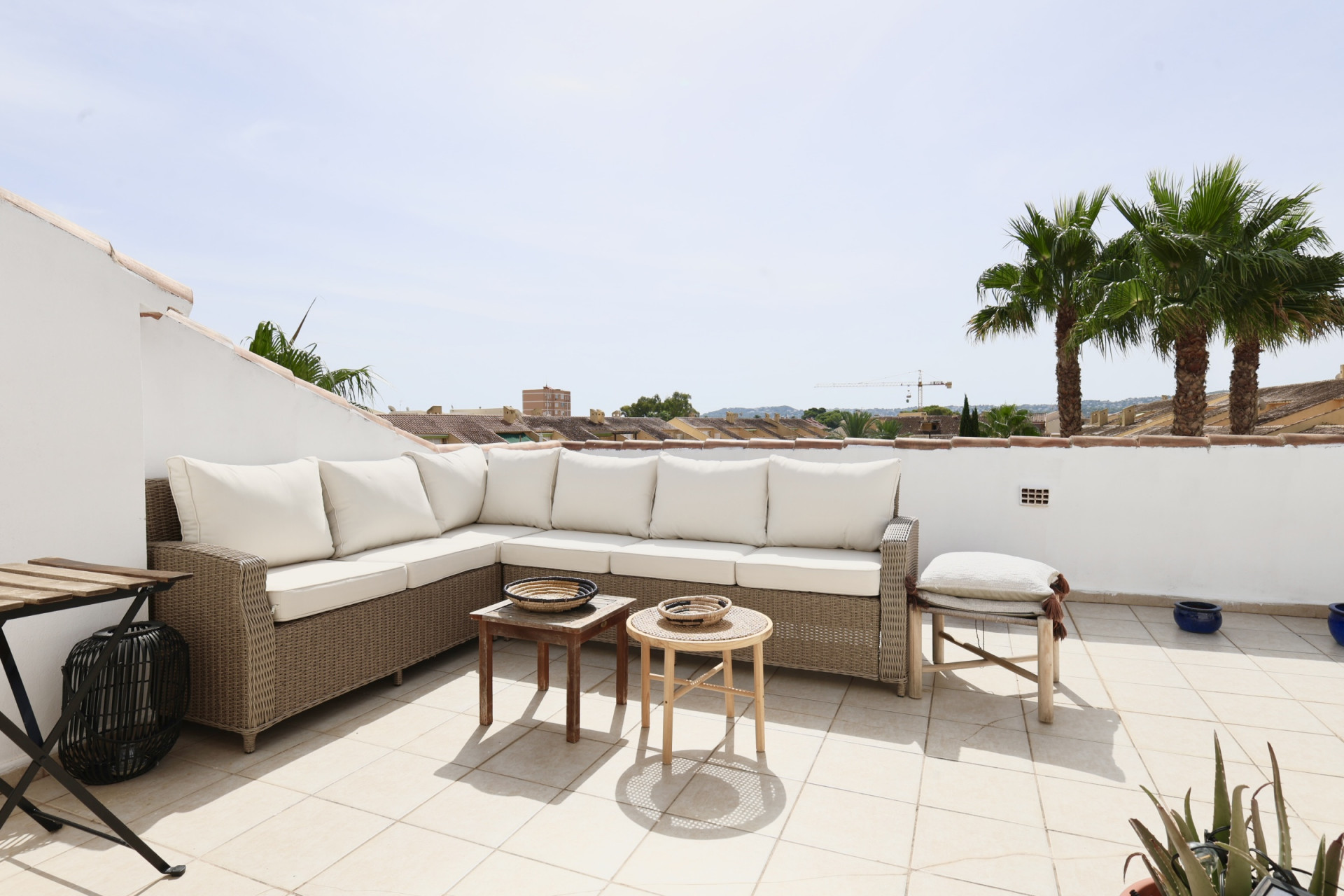 Terrasse mit Korbsitzgruppe und weißen Kissen, mit Blick auf Palmen und Dächer.