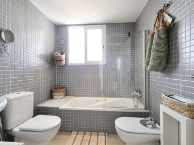 Badezimmer mit grauen Fliesenwänden, Badewanne mit Duschwand und dekorativen Elementen aus Korbweide.