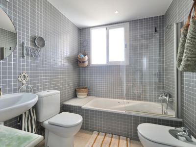 Badezimmer mit grauen quadratischen Fliesen, Waschtisch, Toilette und Bidet. Unter einem Fenster befindet sich eine weiße Badewanne.