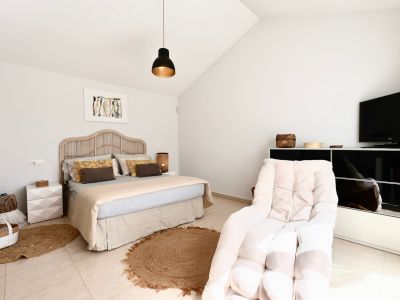 Helles Schlafzimmer mit Rattanbett, Jute-Teppich und Sessel neben einem schwarzen TV-Schrank.